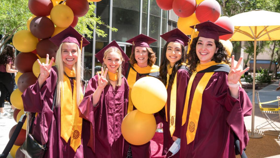 5 smiling women dressed in ASU maroon graduation caps and gowns gather for a photo by a backdrop of maroon and gold balloons.