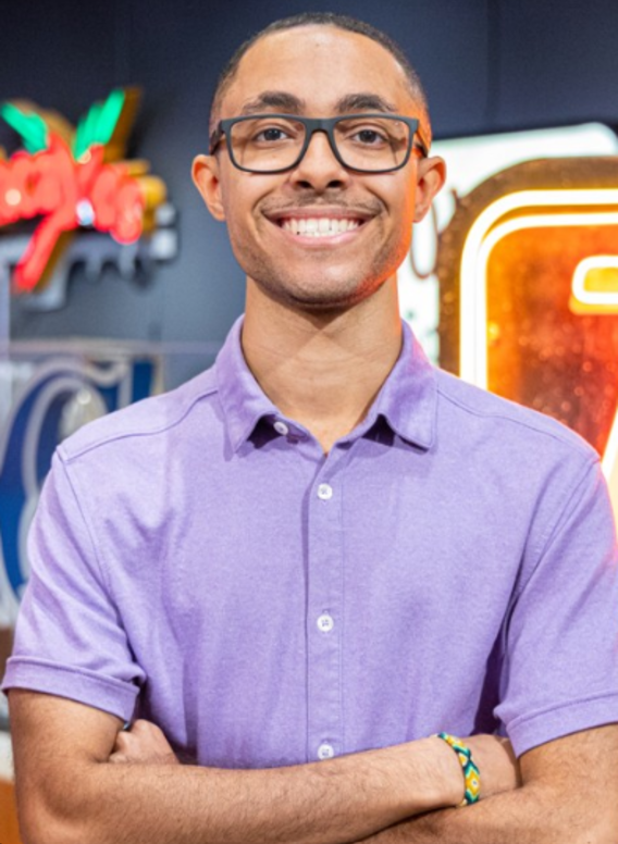 Headshot of Aubrey Feyrer. He is wearing a purple button up, crossing his arms and smiling. He has on glasses and is standing in front of one of the signs from the Tempe Signs collection.