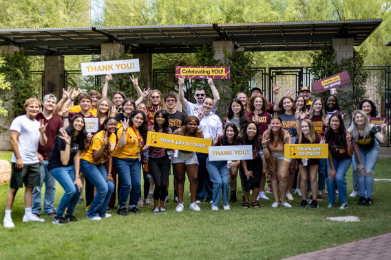 A group of Scholarship recipients all standing together smiling. A few of them are holding signs saying thank you. 