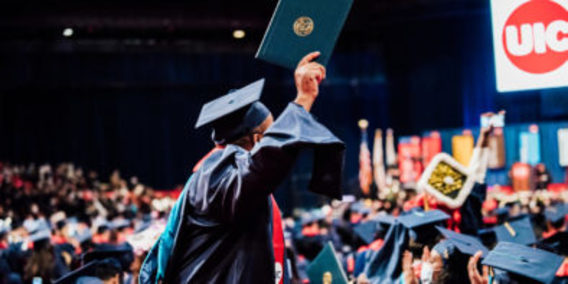 UIC student at graduation ceremony wearing blue commencement gown and red stole. standing and  holding diploma with joy 