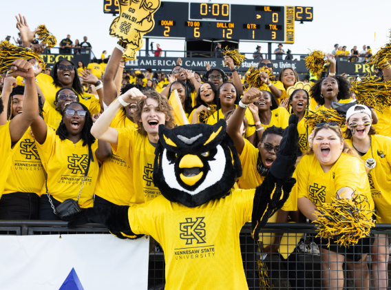 A group of enthusiastic Kennesaw State University students, dressed in yellow, cheer alongside their school mascot at a sports event.