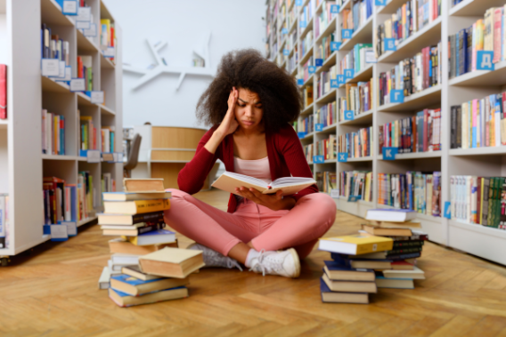 Student sits on the floor of a library, holding an open book and surrounded by stacks of books on the floor. She rests her head on her hand, looking stressed.