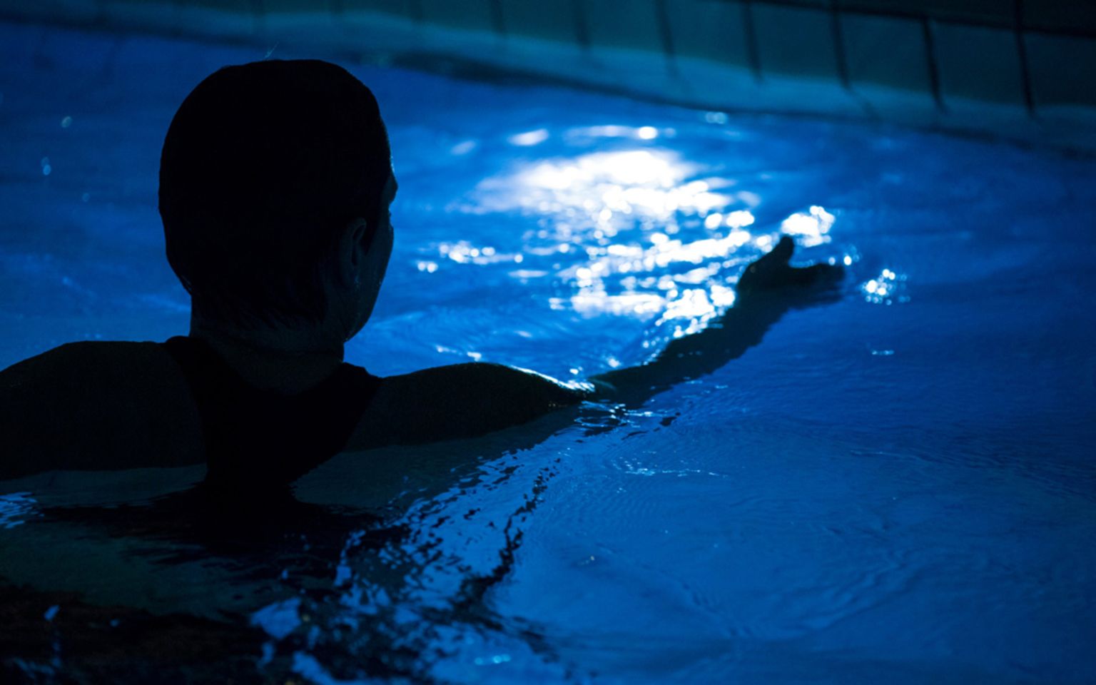 siluett av kvinnas huvud och arm bakifrån i upplyst pool i nedsläckt rum