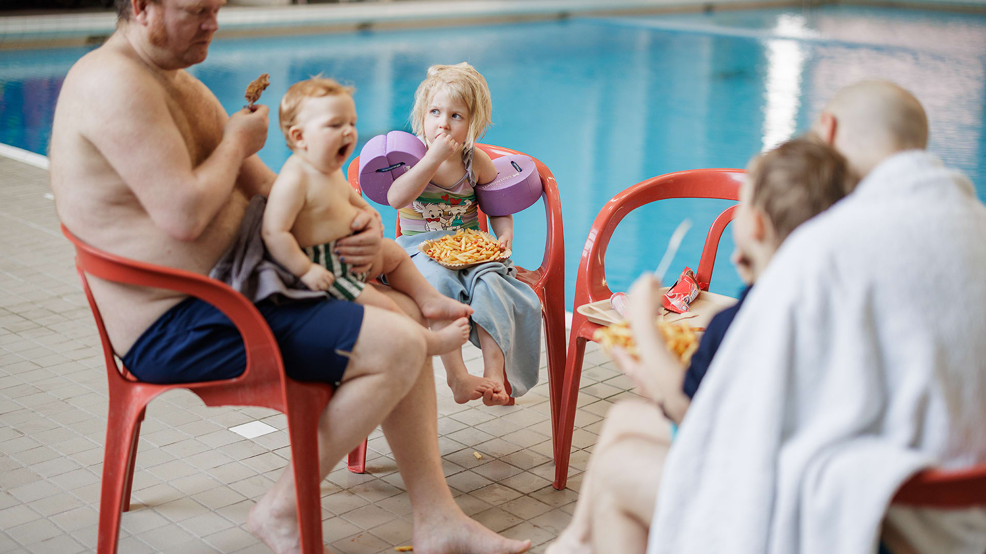 En barnfamilj äter pommes frites vid bassängen i en simhall