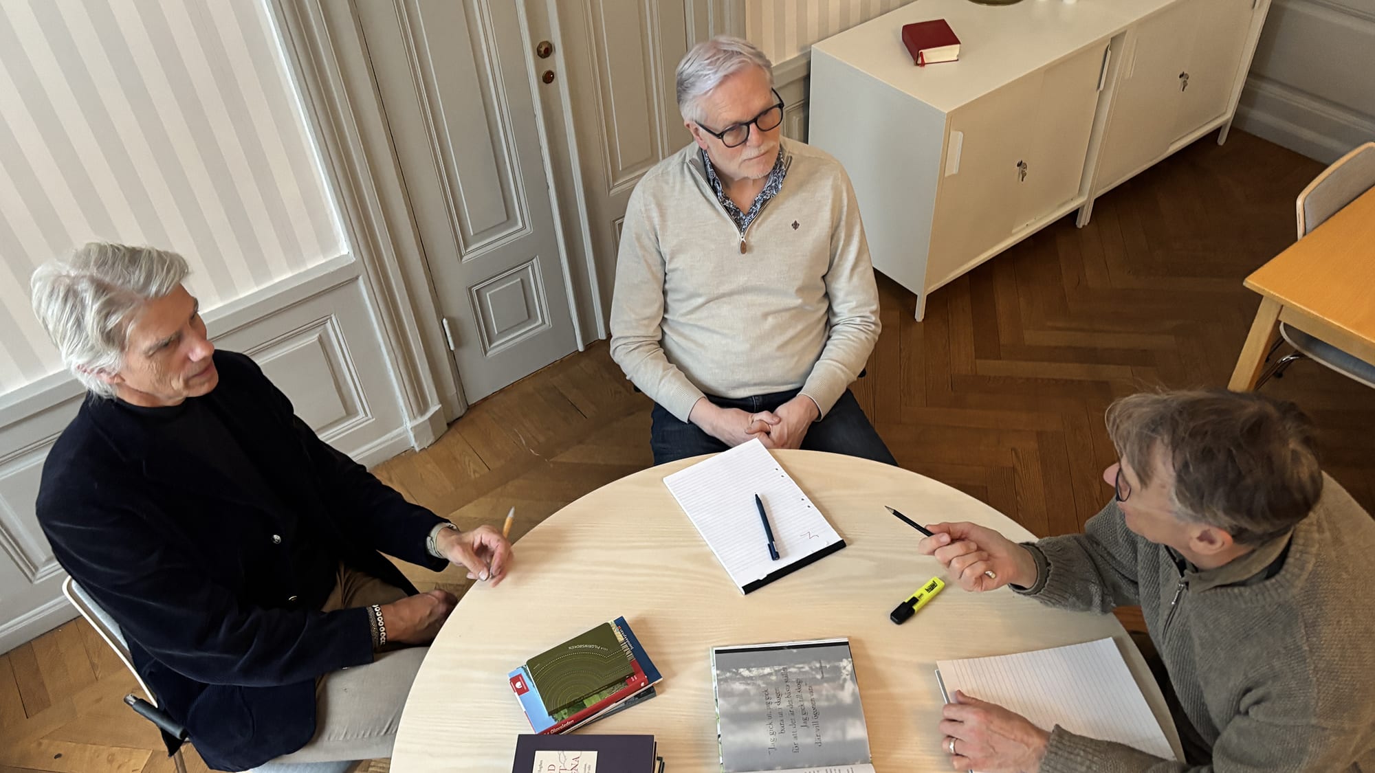 Tre män, Ole i mitten, sitter runt ett bord och diskuterar några böcker. Foto: Sanna Percivall.
