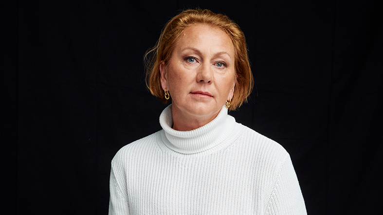 Medelålders kvinna med rak blick i kameran. Vit tröja svart bakgrund