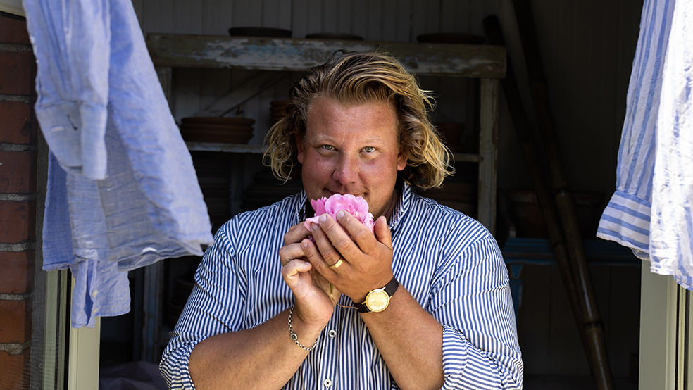 Blond man med page doftar på en rosa ros och tittar in i kameran.