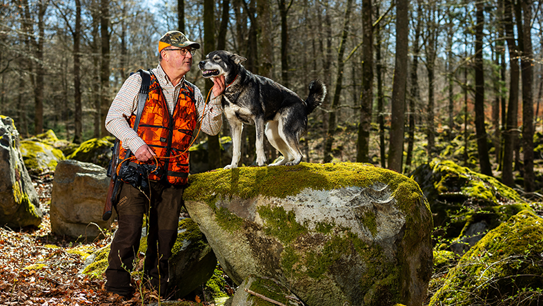 Man står i jaktkläder i skog. Hans hund står på sten bredvid och ska precis få en puss på nosen.