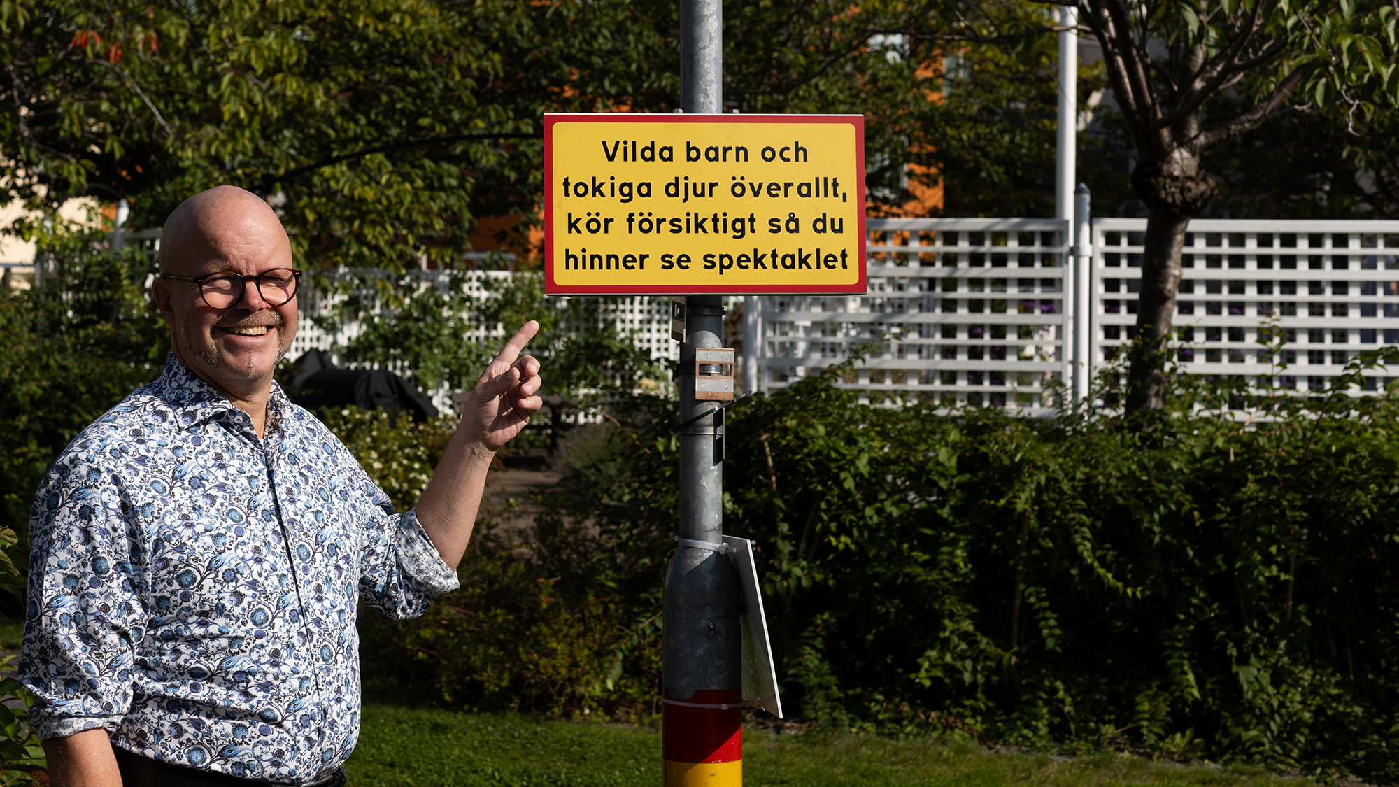 Medelålders man i somrig miljö pekar på en skylt med en rolig text på gatan. 