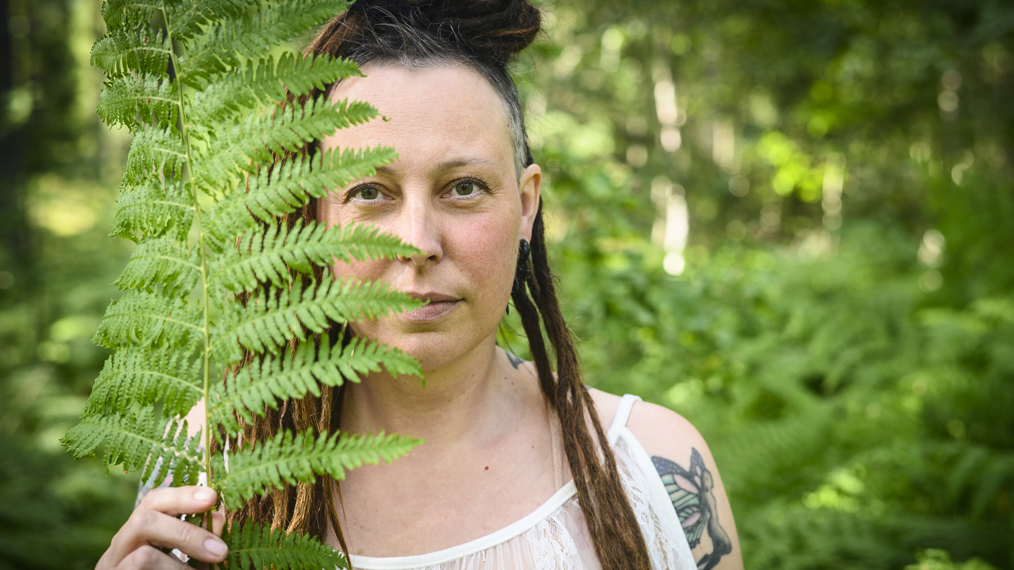 Anna står i skogen och håller upp en ormbunke som delvis täcker hennes ansikte. Fotot är taget av Anna Edlund.