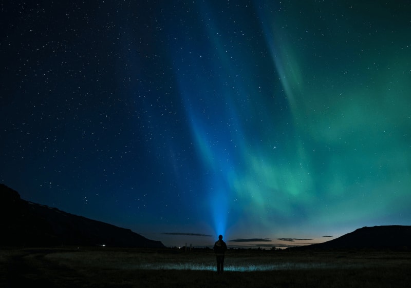 Image of a person standing on the ground in Iceland with a flash light lighting up the sky that is lit by Northern Lights.