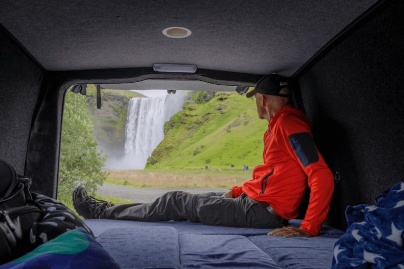 Image of a person sitting inside a campervan looking at a waterfall in Iceland.