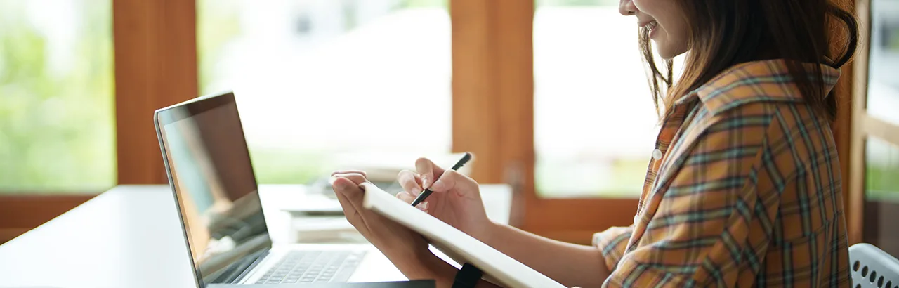 a woman in front of her laptop taking down notes with a pen and notebook