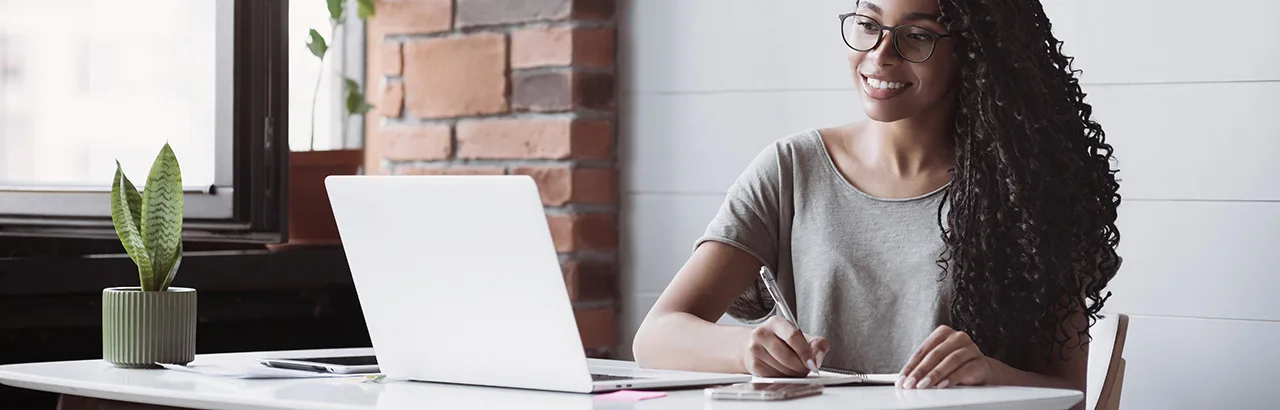 long-haired African-American woman taking online bootcamps, taking notes with a pen and notepad in front of her open laptop