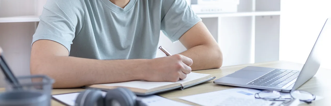 partial image of a man taking notes with a pen a paper in front of an open laptop