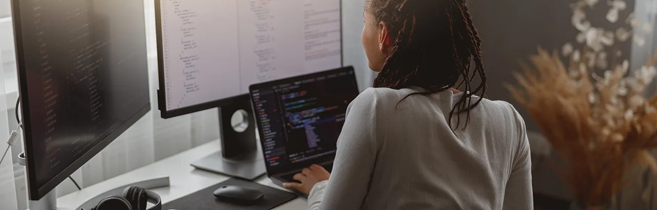 African-American woman working on multiple monitors desktop, studying software engineering via bootcamp online