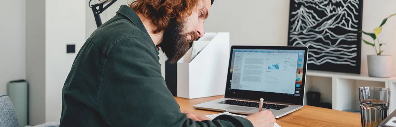 long-haired, bearded man taking notes with a pen and notepad in front of his open laptop while taking an online data analytics bootcamp
