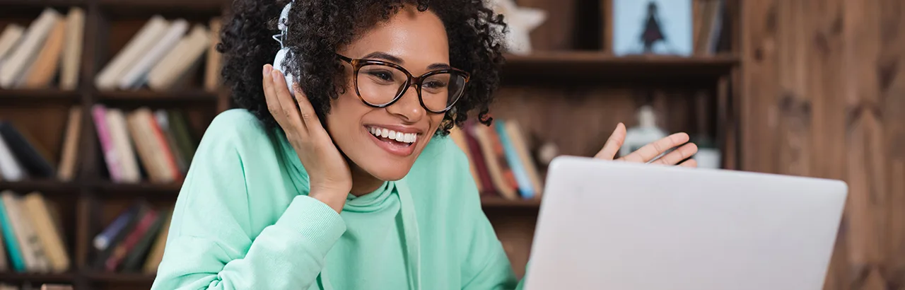 smiling African-American woman in light green sweater and white headphones studying Software Engineering Bootcamp online in front of her laptop