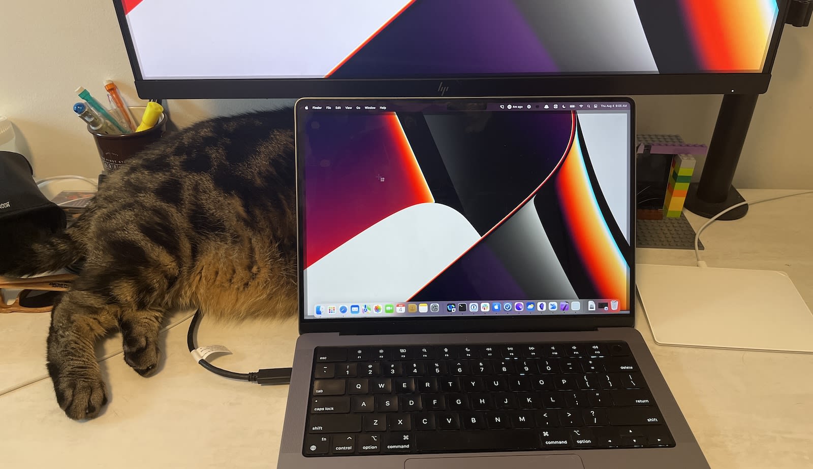 A laptop on a desk, plugged into a bigger monitor, there is a cat sleeping being the laptop but only his bottom half is visible.