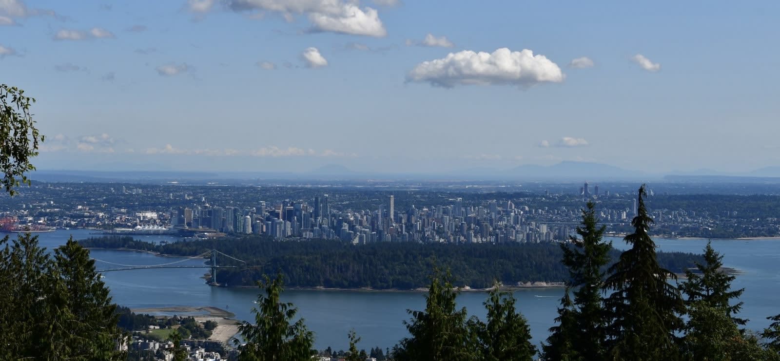 Stunning view of Vancouver, Canada from an elevated vantage point. The foreground features evergreen trees, while the middle ground showcases the city's skyline with its numerous skyscrapers and high-rise buildings clustered near the waterfront. A large suspension bridge, likely the Lions Gate Bridge, connects the mainland to a forested peninsula. The scene is framed by the expansive waters of Vancouver Harbor and English Bay, with distant mountains visible on the horizon under a partly cloudy blue sky.
