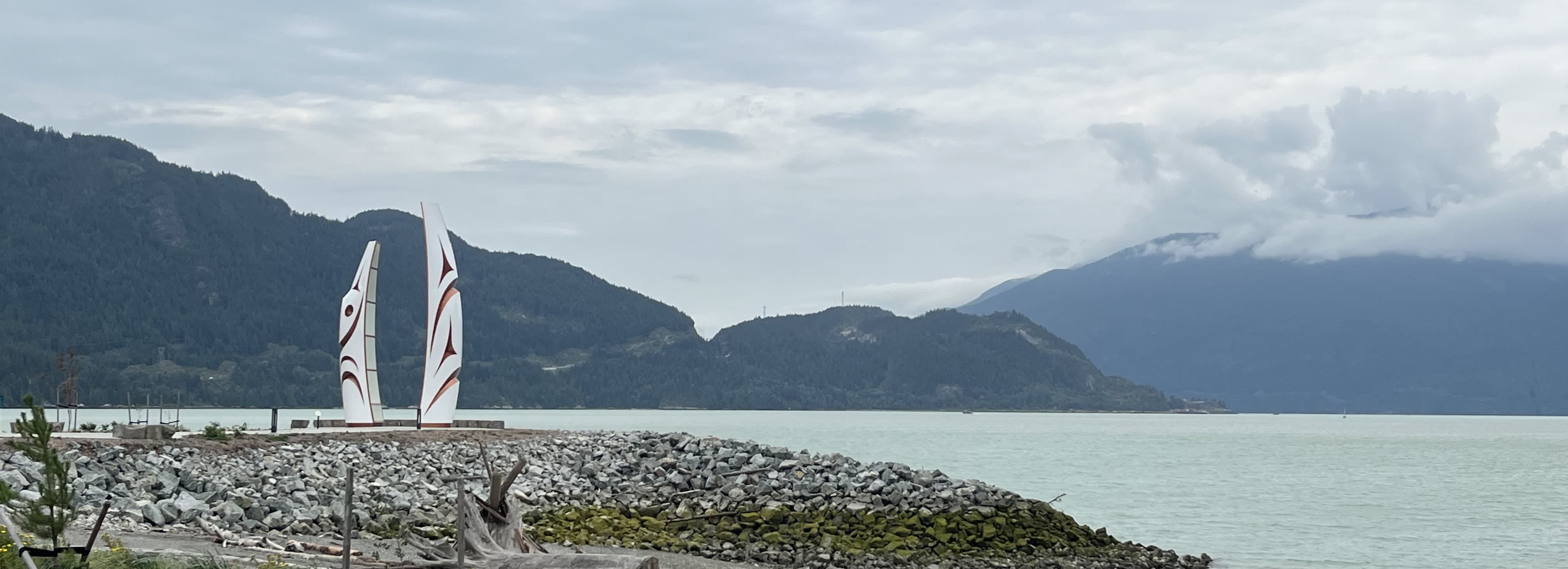 This image shows a serene coastal scene with two tall, stylized sculptures resembling sail-like structures placed on a rocky shoreline. The sculptures feature bold, abstract designs in shades of white and red. Behind them, calm waters stretch toward a mountainous landscape partially obscured by low-hanging clouds. The overall atmosphere is tranquil, with muted colors from the overcast sky enhancing the peacefulness of the natural setting.
