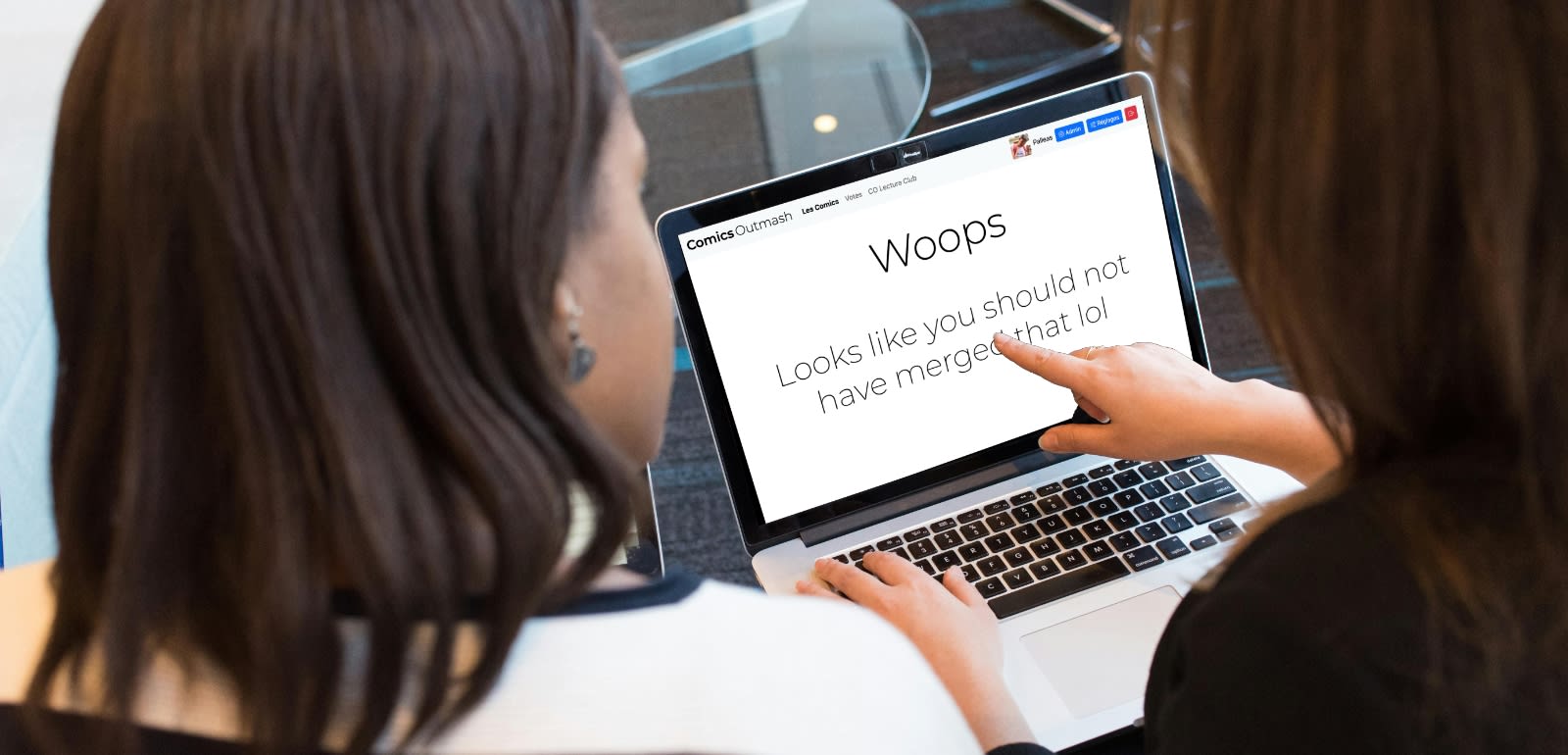 Two people with long hair at a desk, both looking at a laptop's screen. The one on the left is pointing at the screen which shows a page from Comics Outmash that says 
