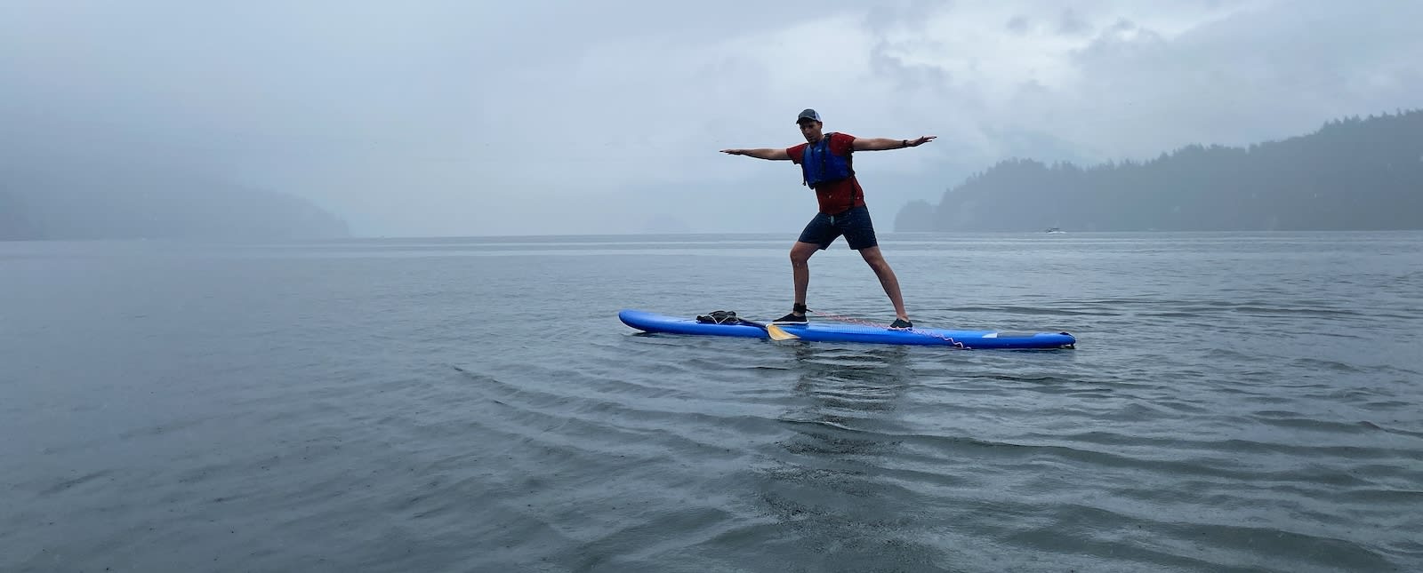 A picture of a me, a man wearing black shorts, a red T-shirt and a blue life jacket. I'm standing on a paddle in the middle of a body of water. In the background is a grey sky, some clouds and mountains.