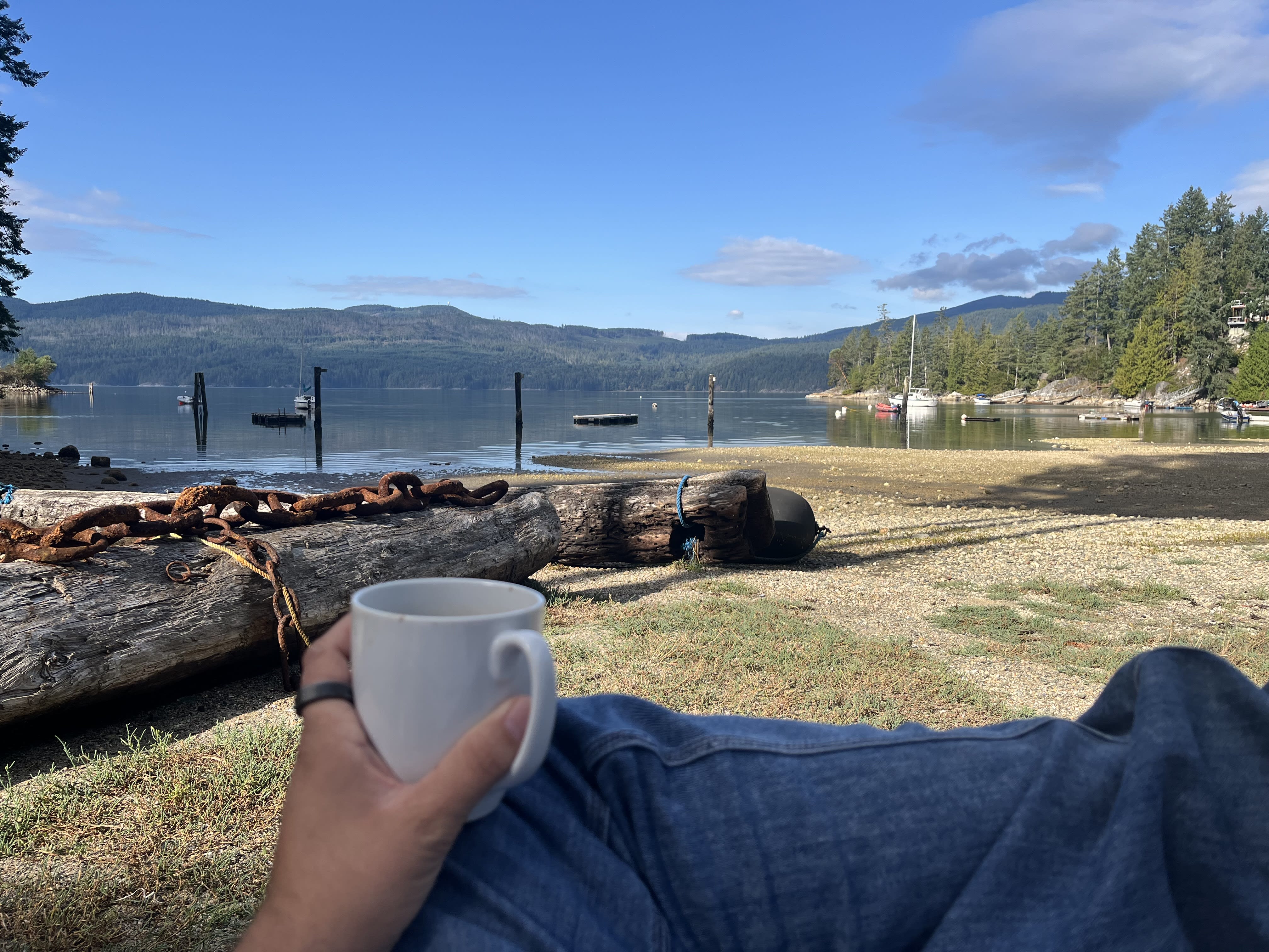 A hand holding a cup of coffee resting on a folded leg. In the background, a beach with stranded logs and a huge rusted chair, the sea with a few boats, the mountains covered with trees and a crisp blue sky.