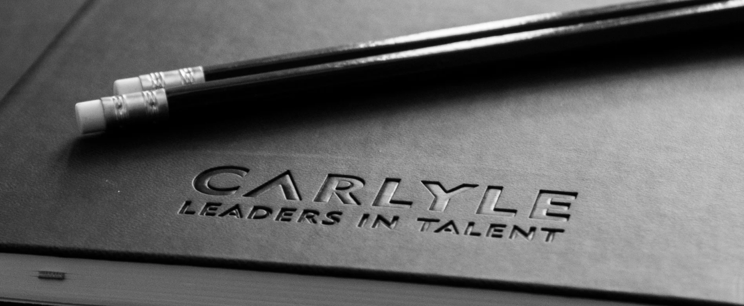 A black notebook with the words "Carlyle Leaders in Talent" engraved into it. Two black pencils have been placed on top of the notebook.
