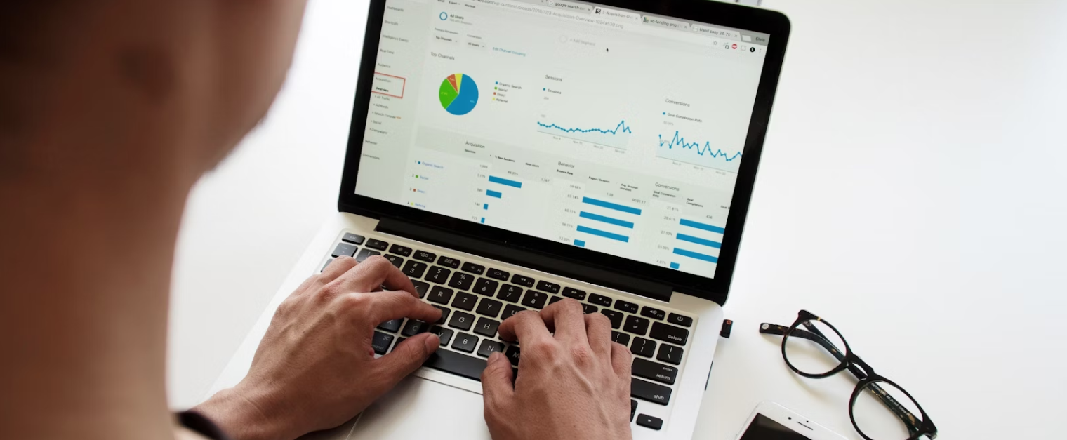 Someone is typing on a laptop while looking at graphs and tables. A phone and glasses are placed next to the laptop.