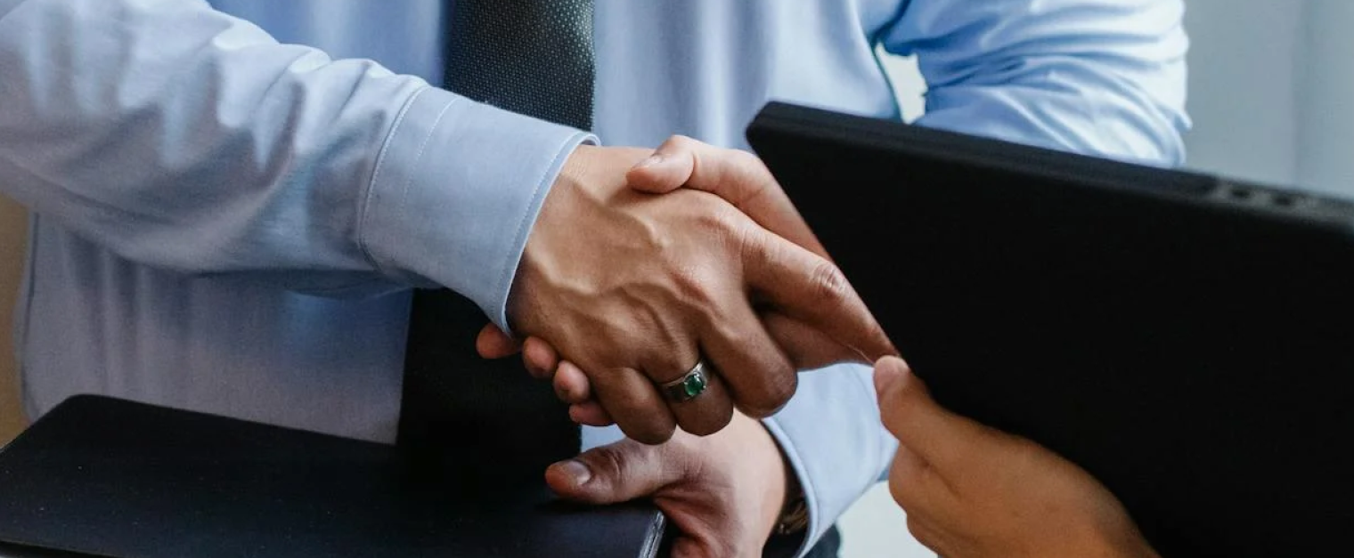 Two people are shaking hands. One is holding a laptop while the other appears to hold a notebook.