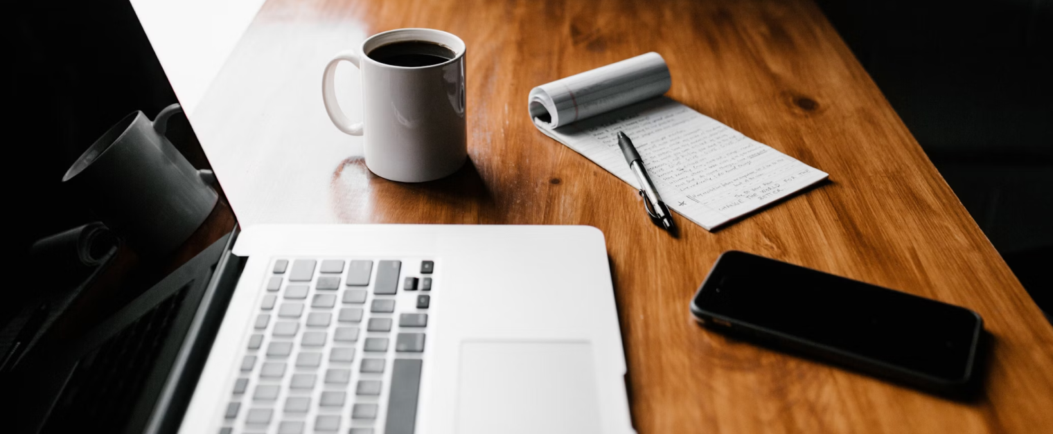 A laptop is open on a desk. There is a coffee mug, notebook and pen placed next to the laptop.