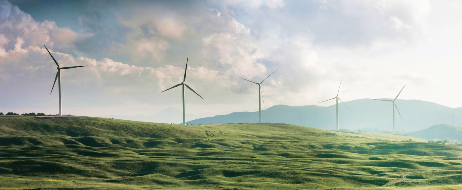 Five windfarms can be seen on top of a green hill. Clouds and a blue sky are in the background.