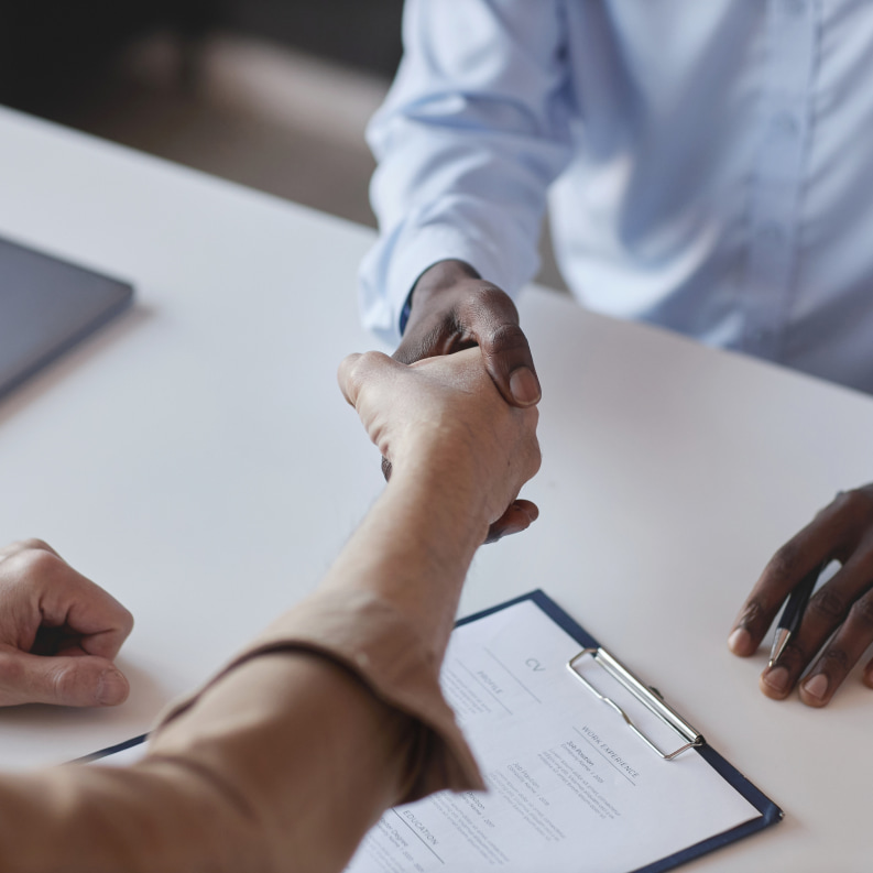 Two people shaking hands while sitting at a desk. A clipboard with a CV has been placed on the desk.