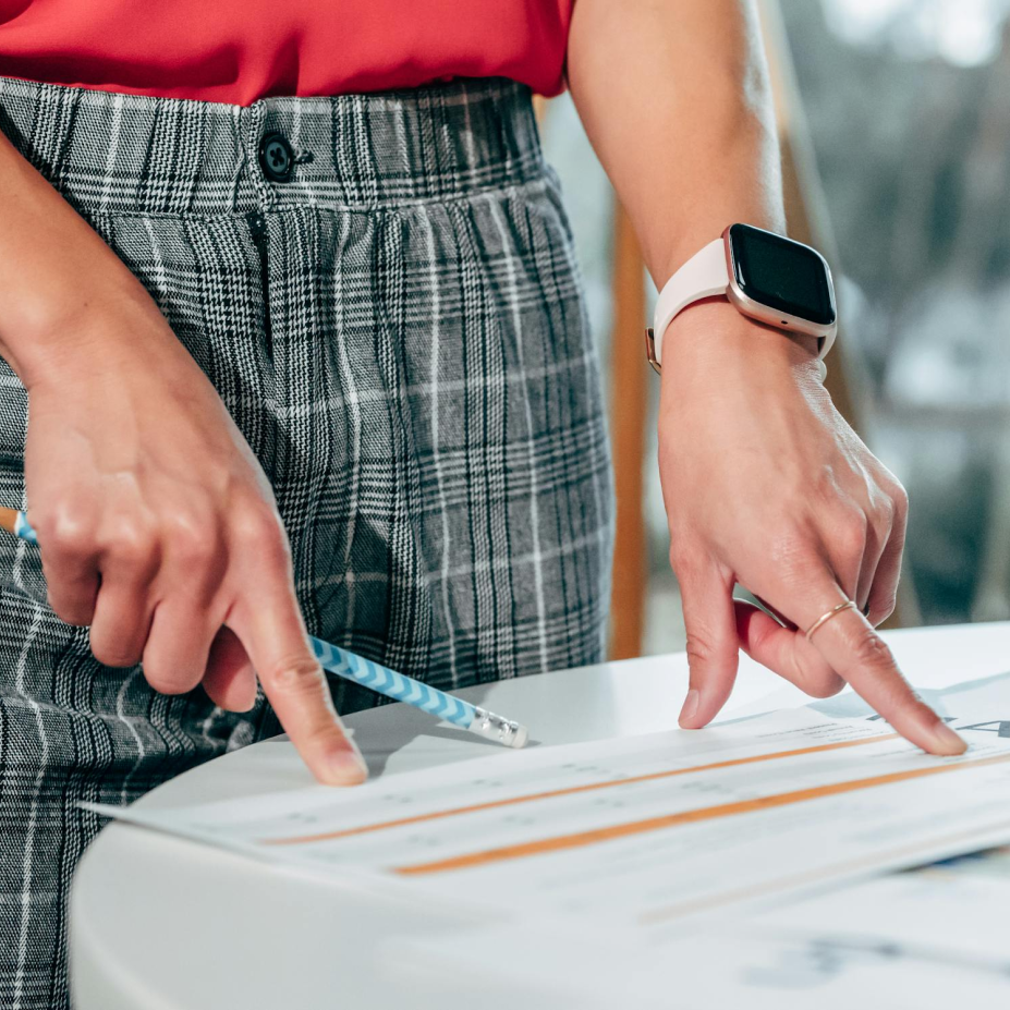 Someone is pointing to a piece of paper on top of a desk. They are holding a pencil in their right hand.