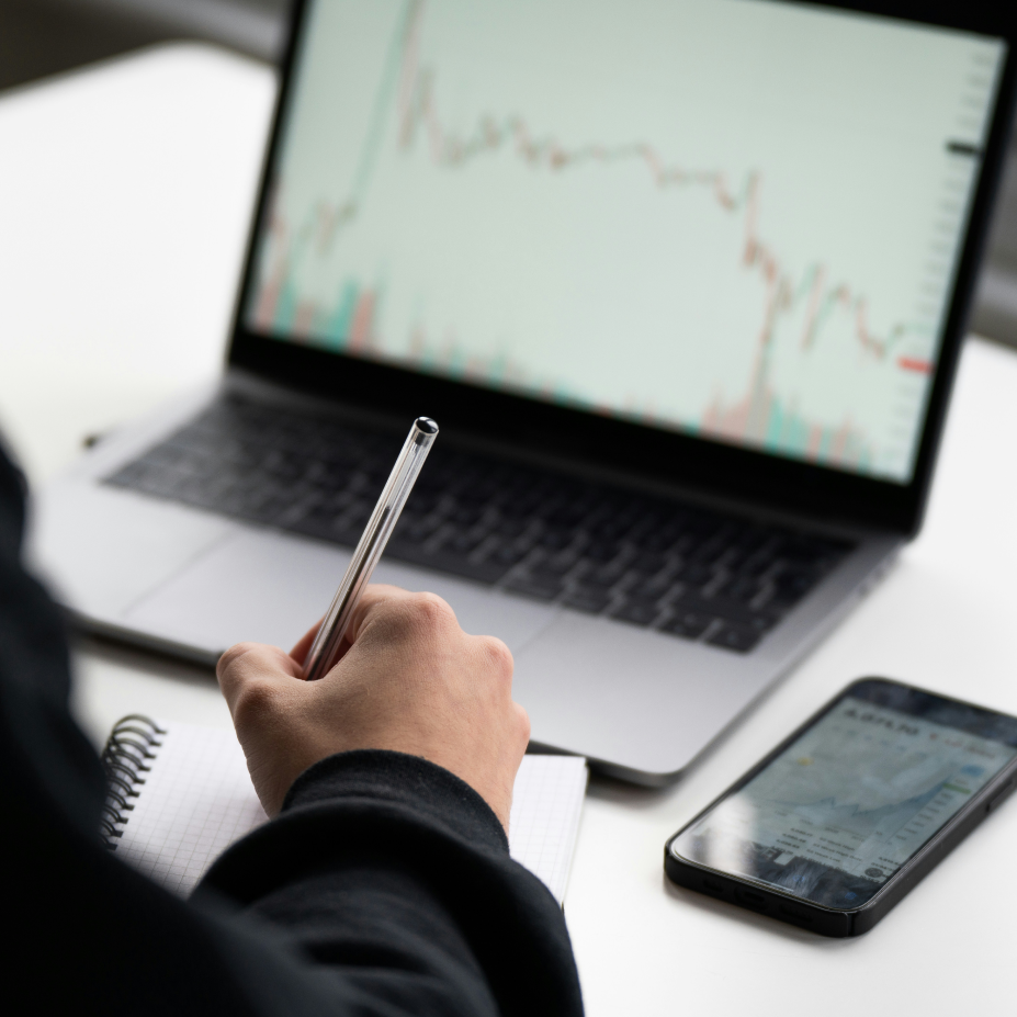 A person is making notes on a notepad at a desk. In front of them is a laptop with a graph displayed. Next to the laptop they have placed a phone on top of the table.