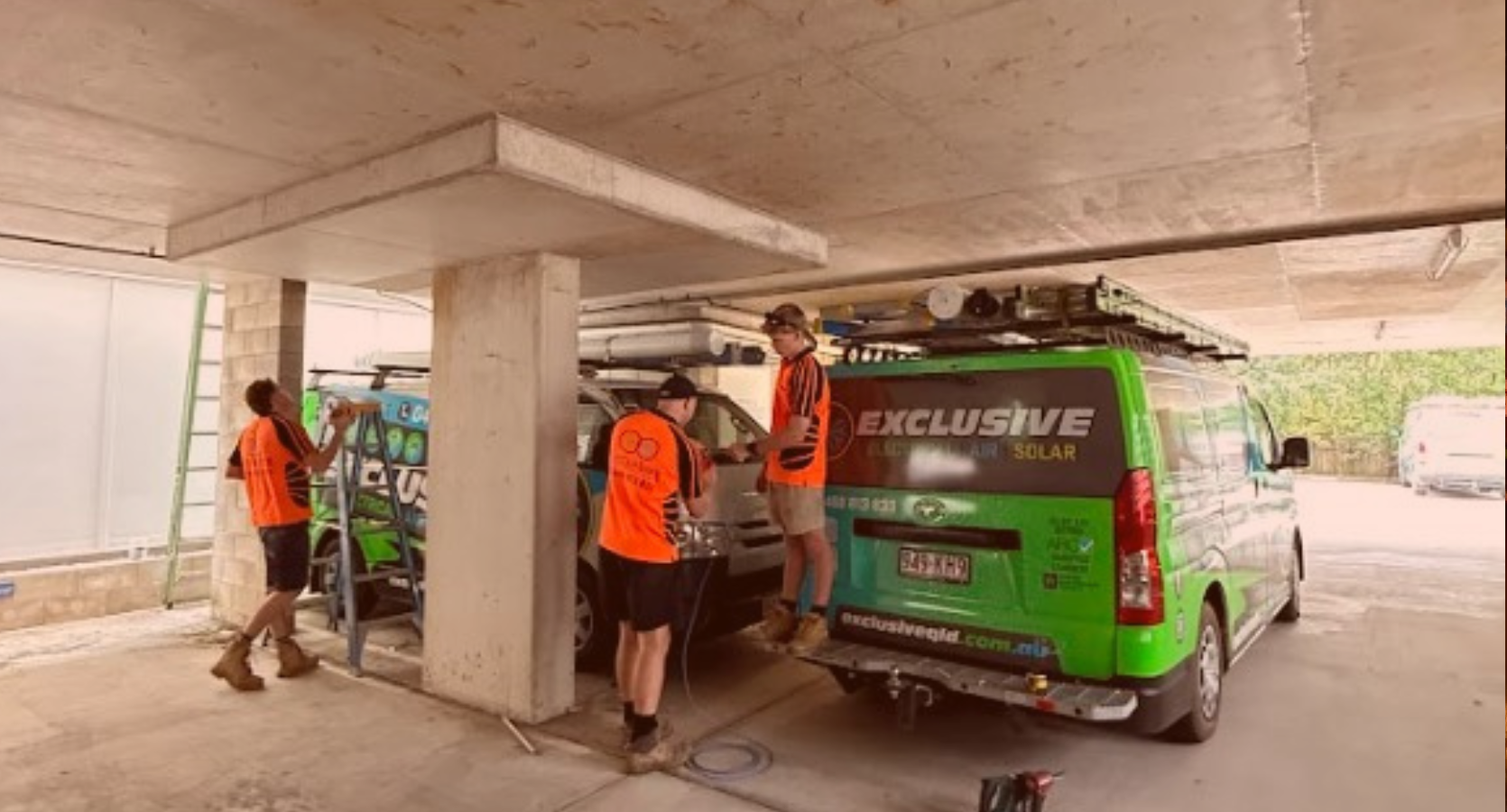 Electricians from Exclusive Electrical & Air in orange uniforms preparing tools next to branded service vans in an underground car park in Brisbane.