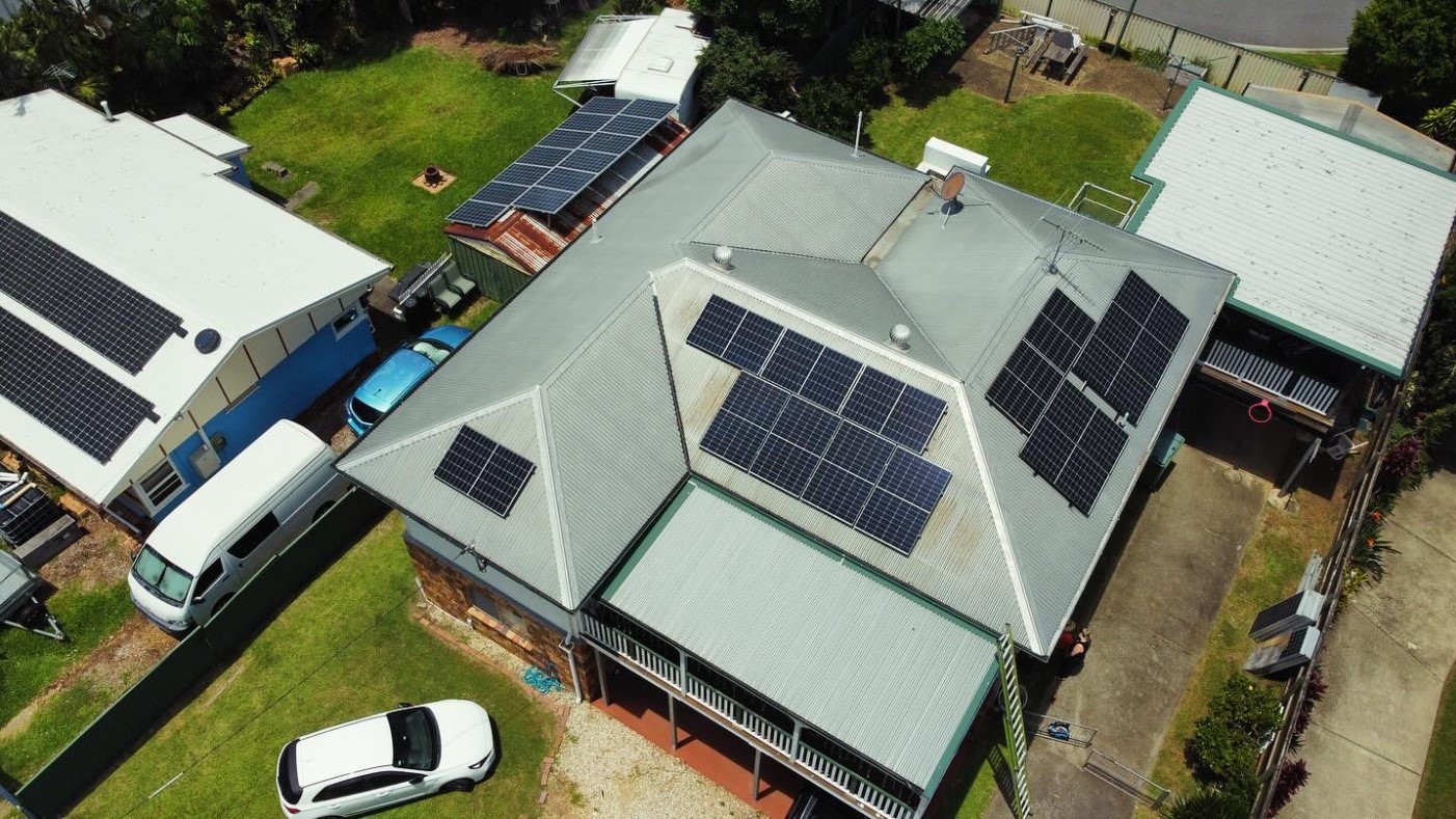 Aerial view of a Brisbane home with multiple solar panel arrays installed on a corrugated metal roof by Exclusive Electrical & Air, showing surrounding sheds and vehicles.