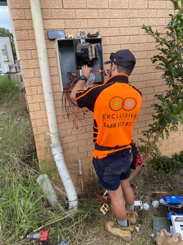 An electrician from Exclusive Electrical & Air working on a switchboard upgrade outside a brick house in Brisbane, wearing the company’s orange and black uniform.