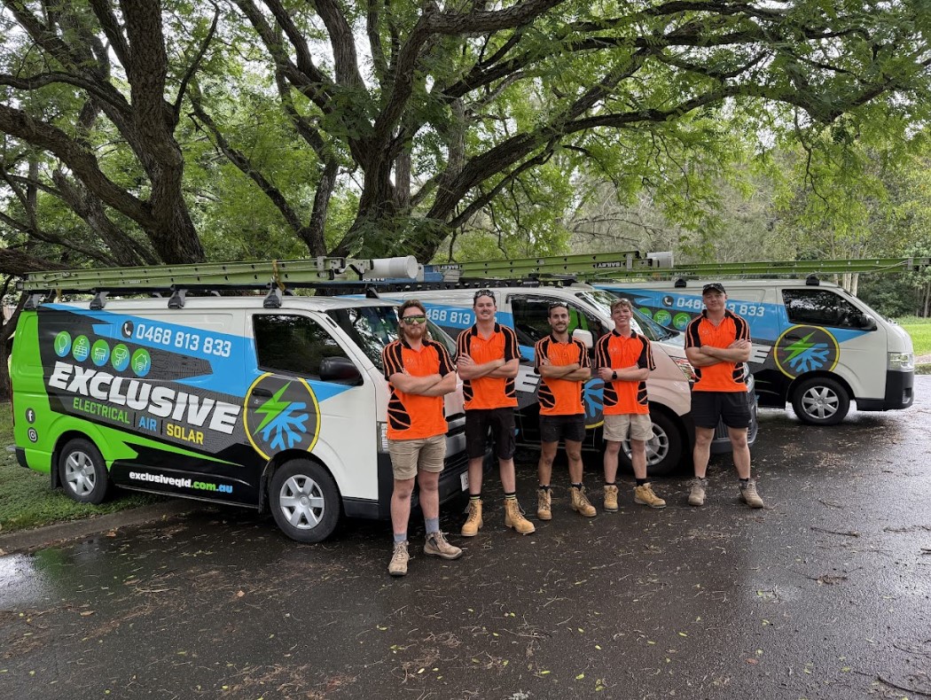 A group of Exclusive Electrical & Air electricians in branded uniforms standing proudly in front of company service vans under large shady trees, showcasing their professional team and fleet.
