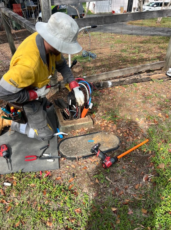 An electrician in safety gear working on underground electrical cables inside a roadside pit, surrounded by tools and equipment during maintenance.