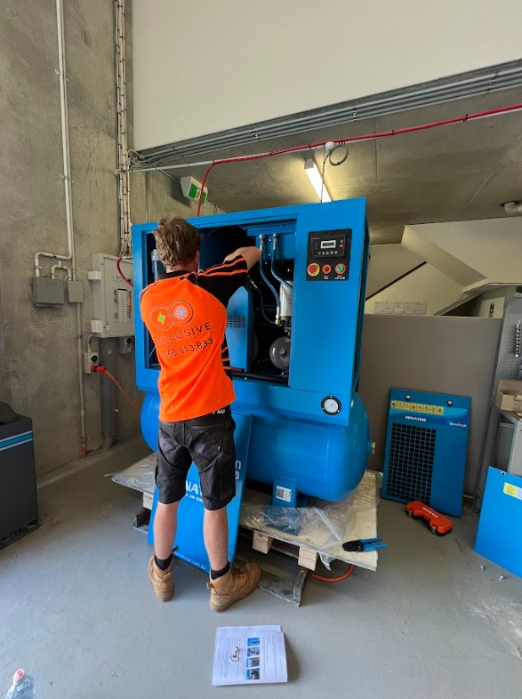 Technician from Exclusive Electrical & Air installing and wiring industrial blue compressor equipment in a commercial facility, ensuring safe and reliable operation.