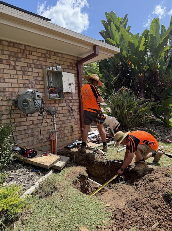 Two electricians from Exclusive Electrical & Air performing trenching work beside a brick house. One is standing near an open electrical meter box, while the other measures inside the trench lined with conduit. Tools and wiring are spread around, with lush tropical plants in the background.