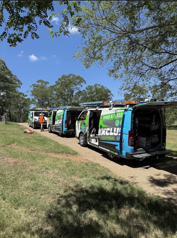 A row of Exclusive Electrical & Air service vans parked along a rural driveway under clear blue skies, with electricians in orange uniforms preparing for a project.