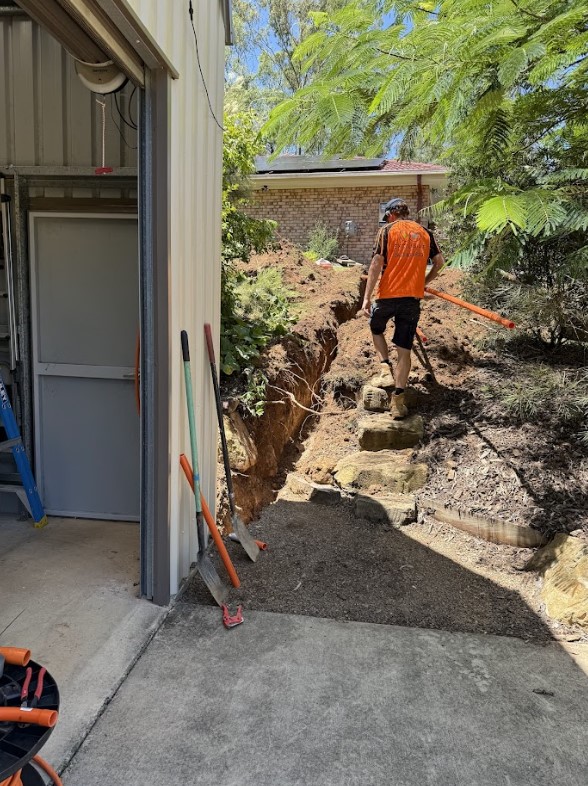 An Exclusive Electrical & Air technician walking up stone steps beside a trench during electrical installation work, with tools and shovels placed near a shed.