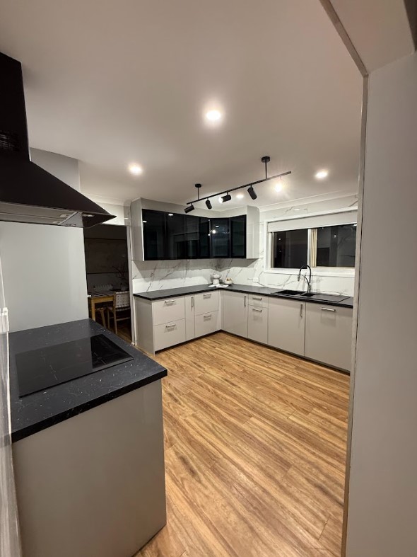 A contemporary kitchen with recessed LED downlights and sleek black track lighting, enhancing the bright and modern design with white cabinetry, black countertops, and wooden flooring.