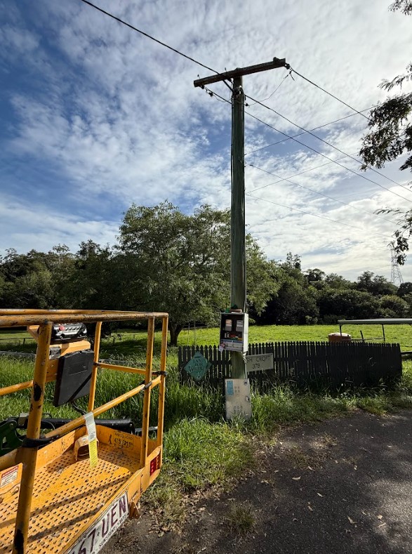 Utility pole with overhead power lines and an outdoor electrical meter box, situated beside a rural property with trees and open field in the background.