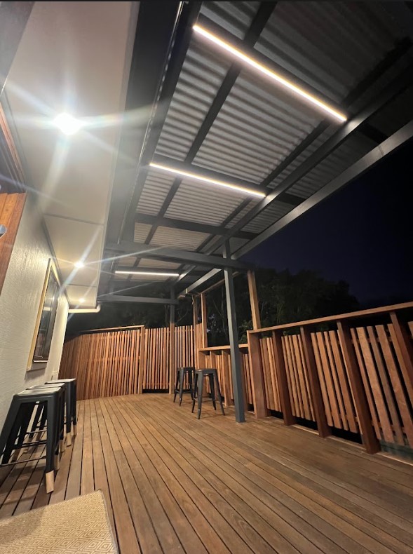 Contemporary outdoor wooden deck with bar stools, timber railing, and stylish LED strip lighting installed under a metal pergola roof, creating a warm and inviting night-time atmosphere.