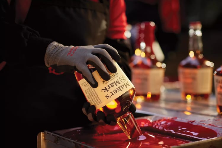 A pair of gloved hands in the left side of the screen are dipping a Maker's Mark bottle, neck down, into a container of hot red wax