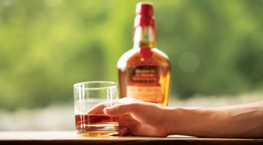 Hand holding chilled glass of Maker's Mark bourbon whisky with blurred background of people sitting at a picnic table